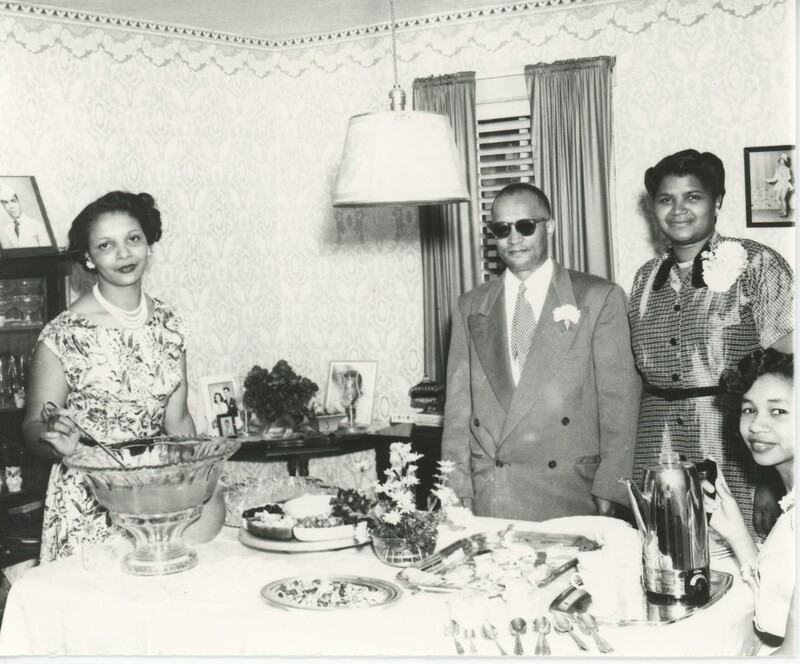 Three people, two of which are standing and one is sitting, are at one side of a food table for a party and one person is stood at the other end by a punch bowl holding the laydle spoon.